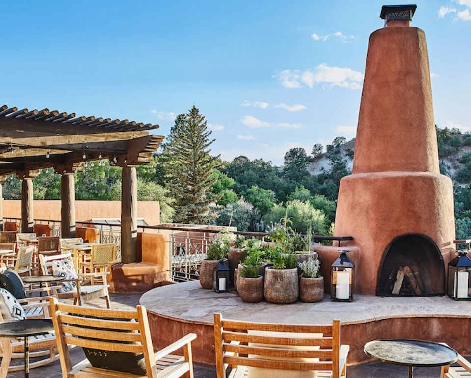 Outdoor seating area with terracotta fireplace and wooden furniture surrounded by greenery, under a clear blue sky.