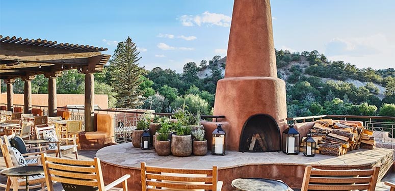 Outdoor terrace with a large adobe-style fireplace, surrounded by wooden chairs and overlooking lush green scenery.